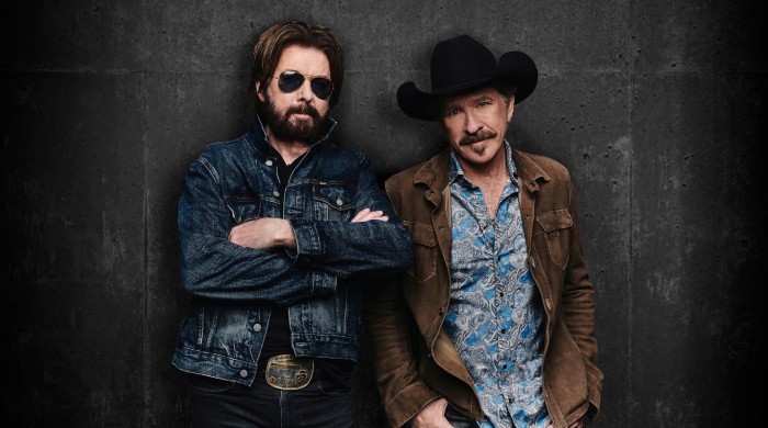 Brooks & Dunn stood against a black wall one wearing a denim jacket and black sunglasses, the other a brown jacket and black cowboy hat